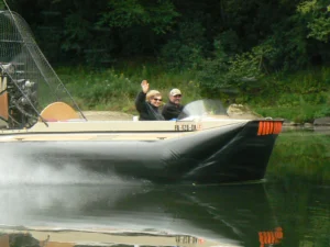 Two people waving from Shamus' Geoduck hovercraft on a river